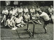 Black and white photo of ladies' hurdles coaching showing a group of female students sat on the grass watching a female academic jump a hurdle.